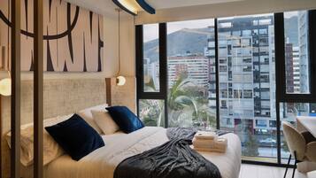 Ein modernes Hotelzimmer mit einem großen Bett, einem Schreibtisch und Blick auf die Stadt durch große Fenster.