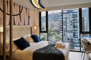 A modern hotel room with a large bed, a desk, and a view of the cityscape through large windows. - Cielo Residences by Nomad-in (Quito)
