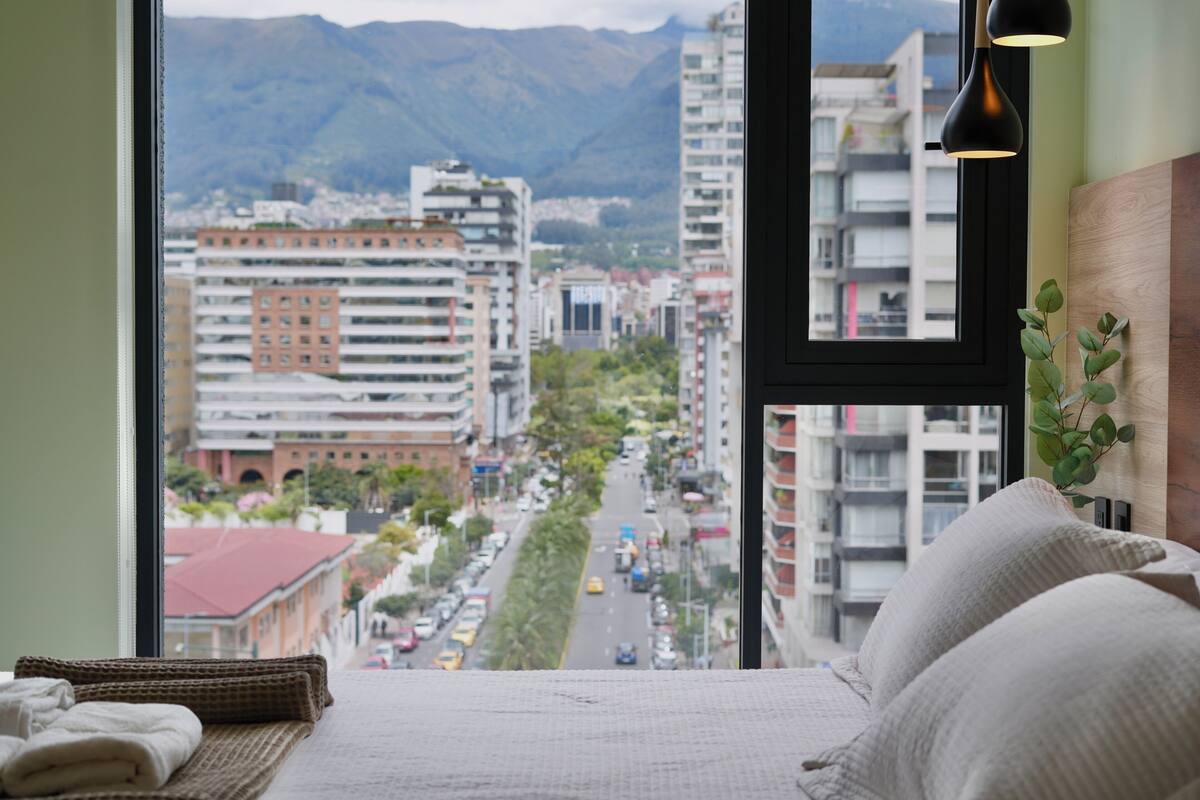 Habitación de hotel con cama, vista a la ciudad y ventana con montaña al fondo.