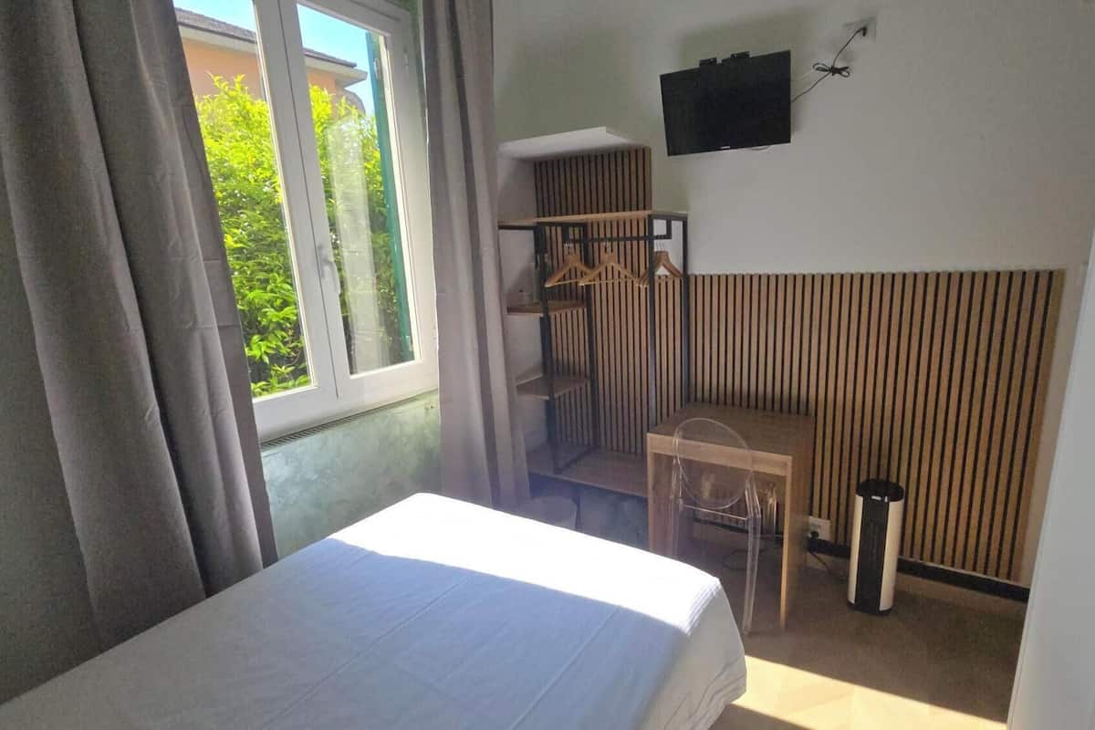 A room with a bed, a small table, a chair, a TV mounted on the wall, and a window with curtains.