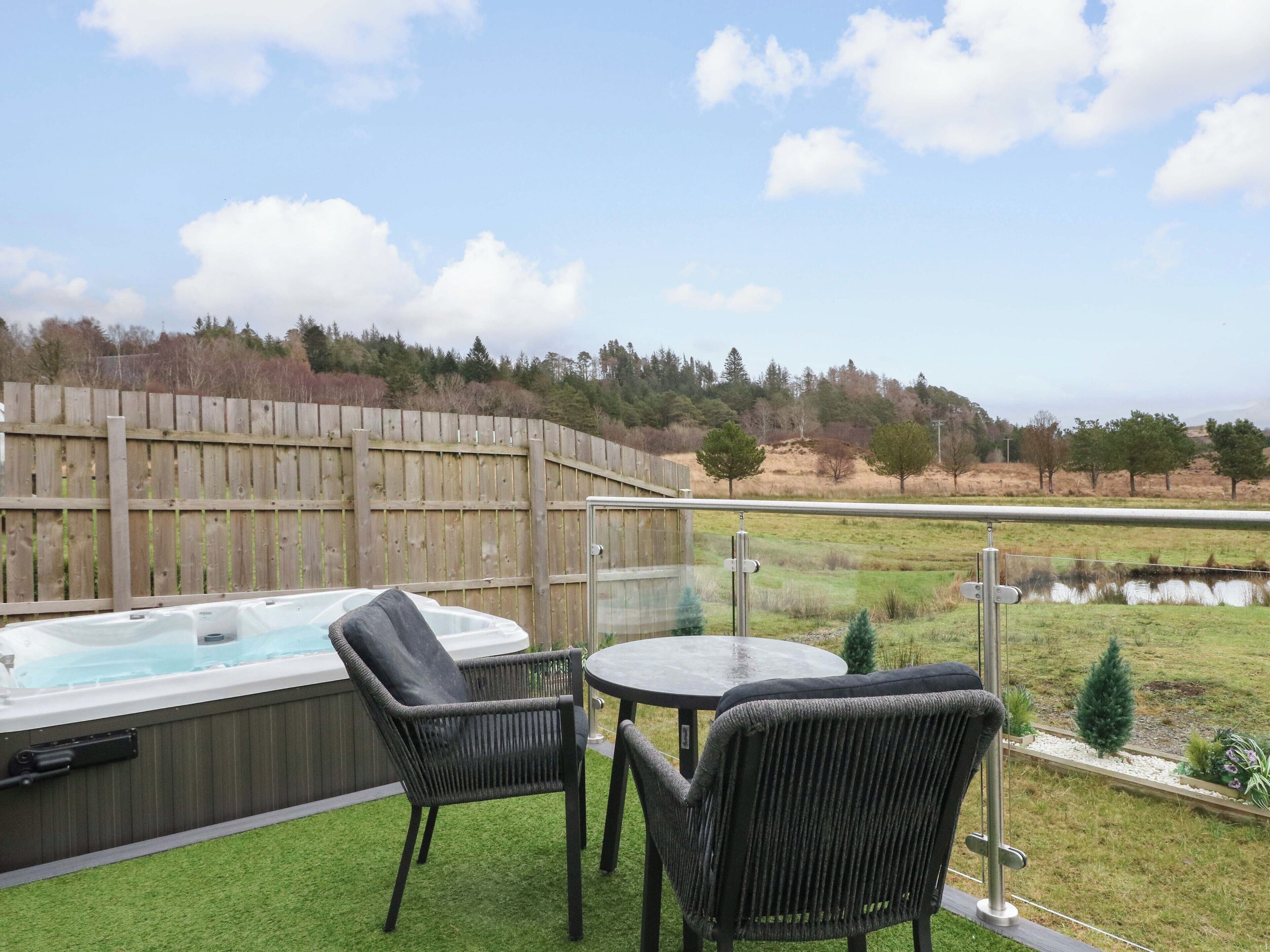 Un jacuzzi con dos sillas y una mesa en un patio que ofrece vistas a un estanque y árboles.