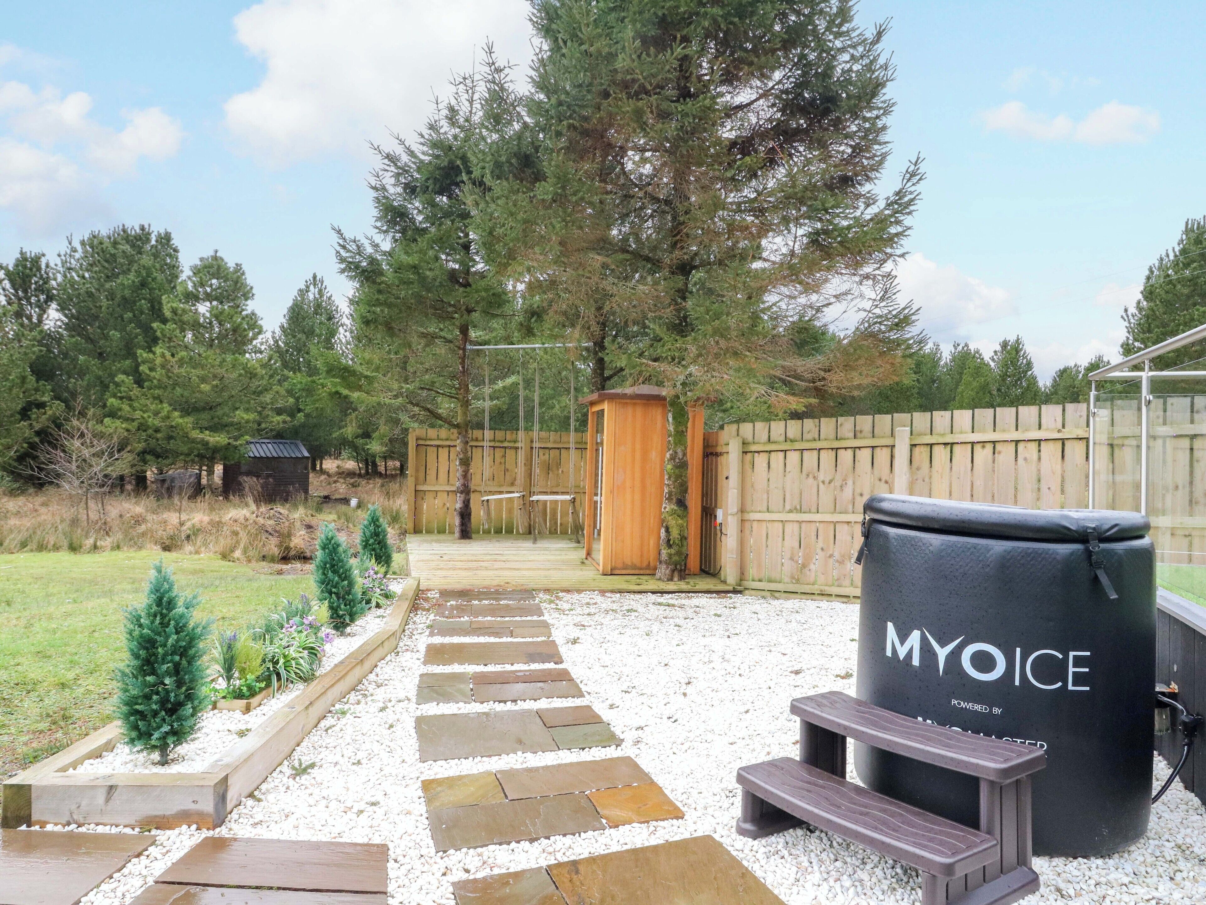 Een achtertuin met een houten terras, een schommelset, een klein houten bouwwerk en een barbecue met de tekst 'MYOICE'.