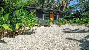 Una casa moderna con vialetto in ghiaia, immersa nel verde di piante e alberi tropicali.