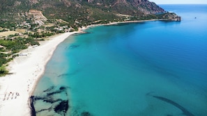 Vista aérea de uma área costeira com uma praia de areia, águas azuis cristalinas e montanhas ao fundo.