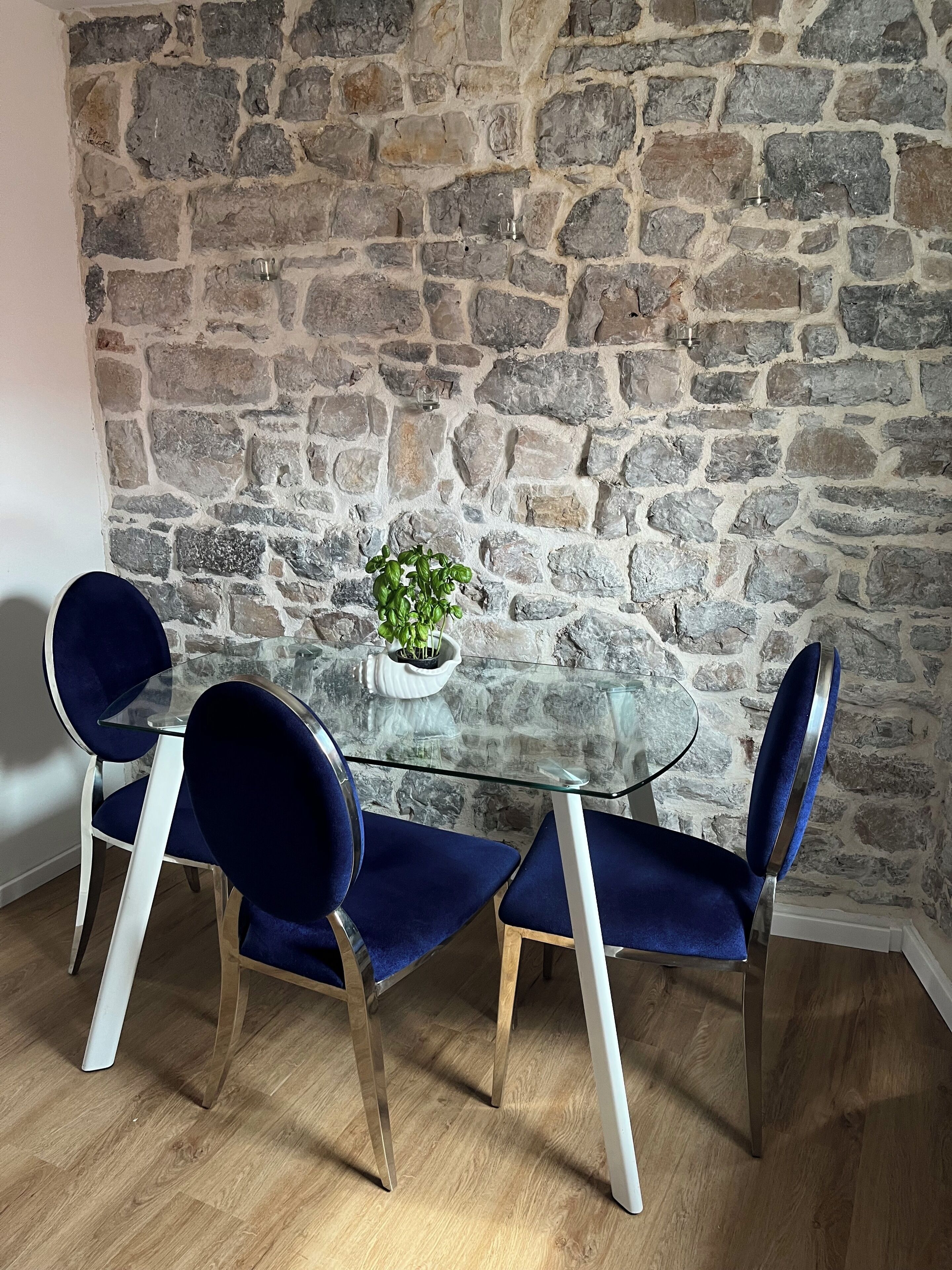 Un área de comedor con una mesa de vidrio rodeada por cuatro sillas azules, una planta en maceta y una pared de piedra.