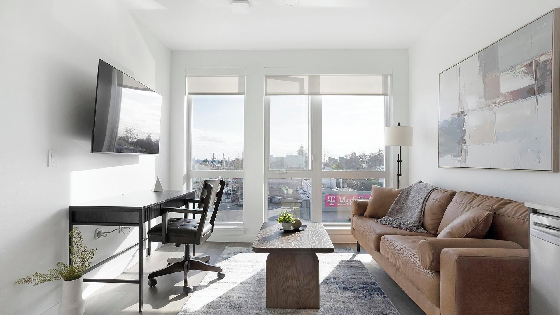 A modern living room with a sofa, a coffee table, a desk, and a TV. There are large windows with a view of the city.