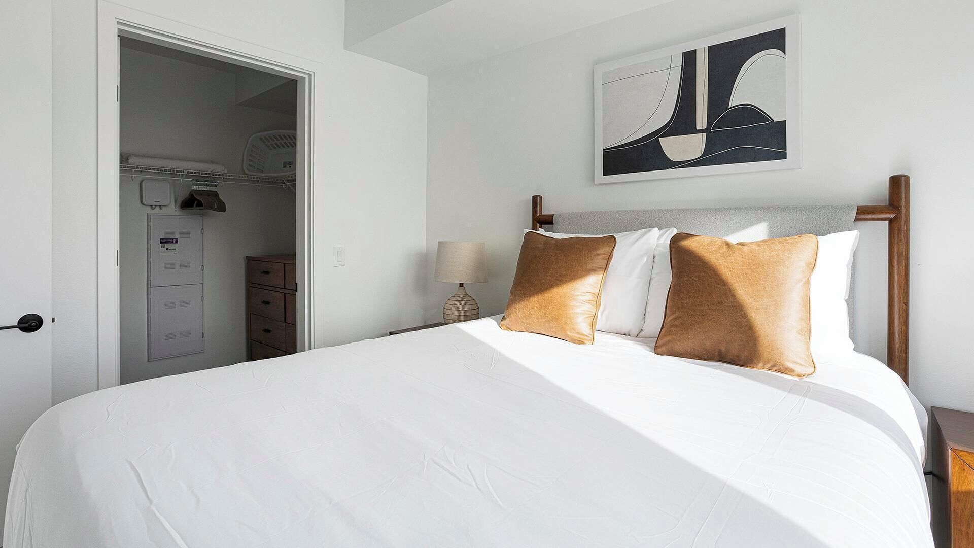 Un lit bien fait, avec du linge de lit blanc et deux oreillers marron, une lampe de chevet et une œuvre d’art abstraque encadrée accrochée au mur.