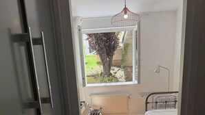 Ein Schlafzimmer mit Bett, Heizkörper, Fenster mit Blick auf Grünfläche und einer Pendelleuchte.