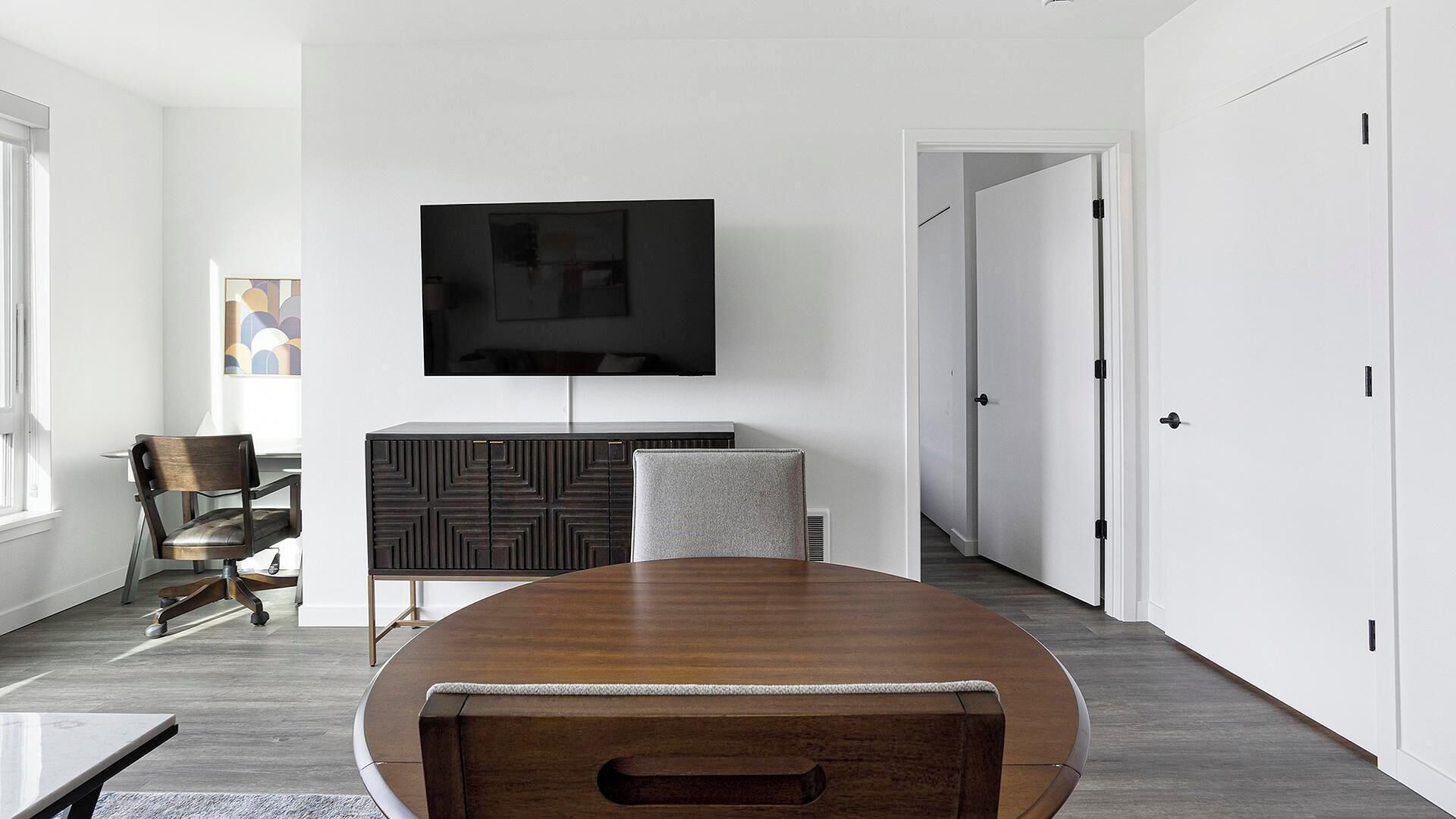 A modern dining room with a wooden table, a TV mounted on the wall, and a cabinet with geometric patterns.