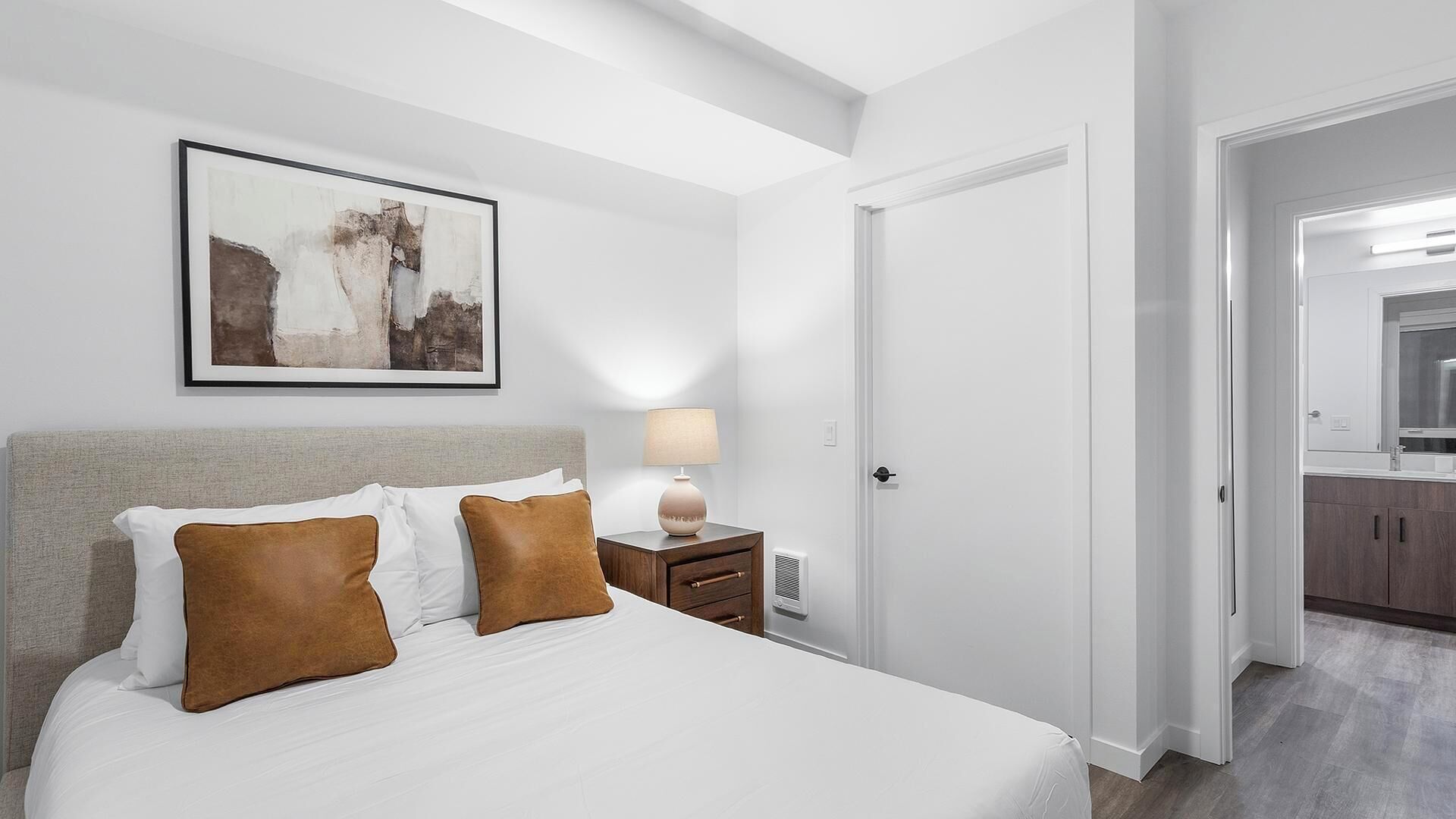 A hotel room with a bed, a nightstand, a lamp, a framed picture, and a door leading to a bathroom.