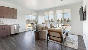 A modern kitchen with wooden cabinets and a built-in oven, a dining area with a table and chairs, and large windows offering a view of trees and buildings. - Landing | Stylish 2BD, Gym (Seattle)