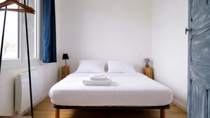 A single bed with white bedding, a wooden floor, a radiator, a wooden nightstand with a lamp, and a window with blue curtains. - House near the sea – sleeps 6 people (Les Sables-d´Olonne)