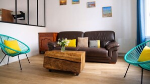 A living room with a brown sofa, two turquoise chairs, a wooden coffee table with flowers, and three framed pictures on the wall. - House near the sea – sleeps 6 people (Les Sables-d´Olonne)