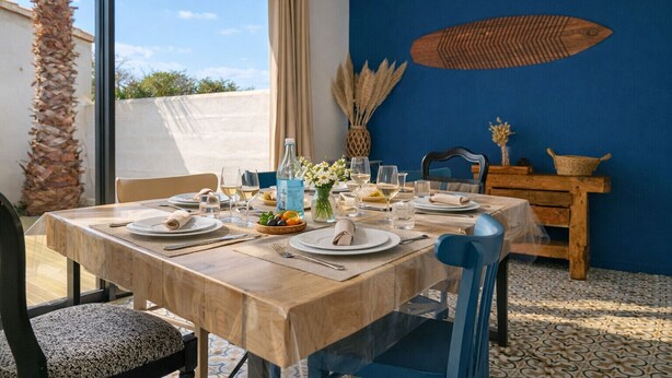 A dining table set with plates, glasses, and a bottle, in a room with a blue wall and a palm tree outside. - House near the sea – sleeps 6 people (Les Sables-d´Olonne)