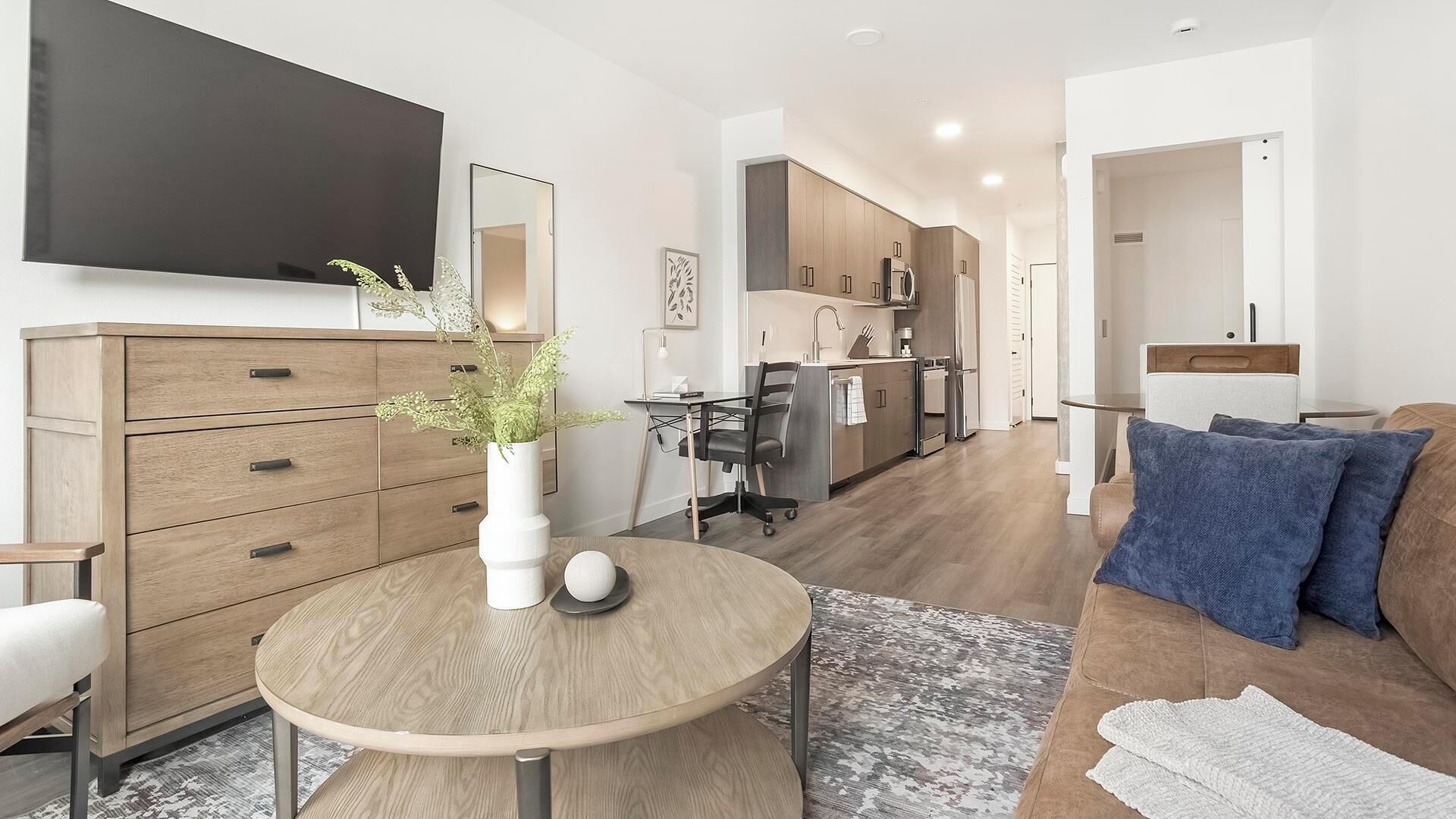 Ein modernes Wohnzimmer mit einem Holzesstisch, einer blauen Sofaausstattung und einem an der Wand montierten Fernseher. Im Hintergrund ist eine Küche zu sehen.