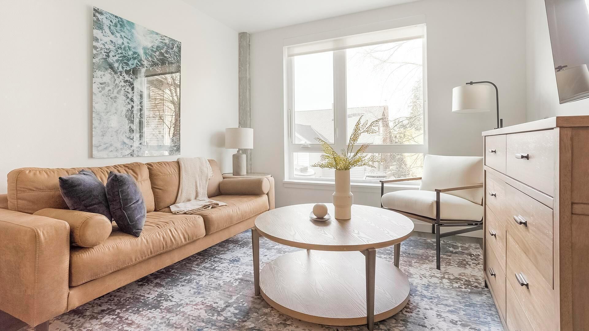 Una sala moderna con un sofá color beige, una mesa de centro redonda de madera y un aparador blanco.