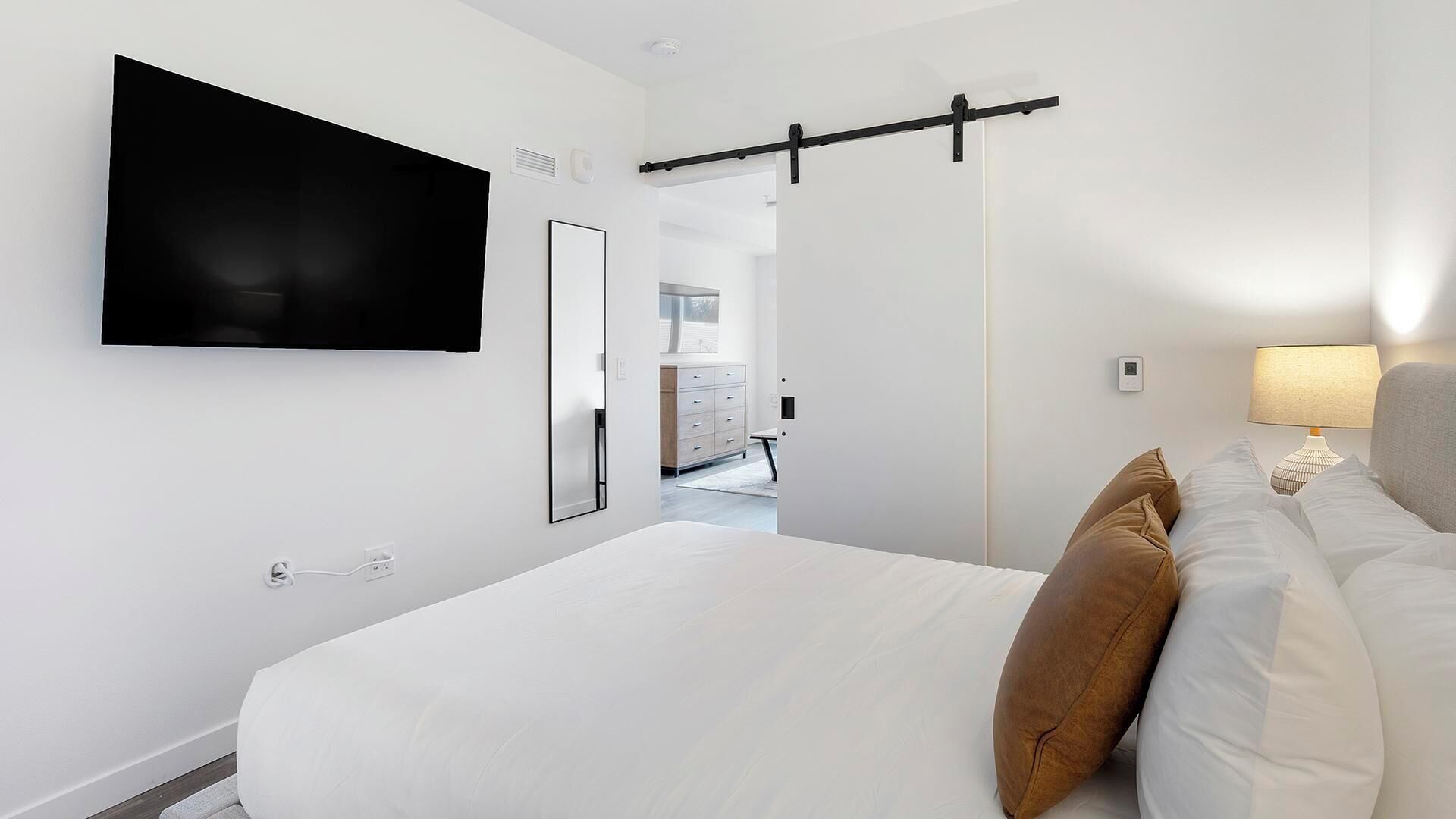 A modern hotel room with a large bed, a mounted TV, a sliding door, a bedside lamp, and a view into the adjacent room.