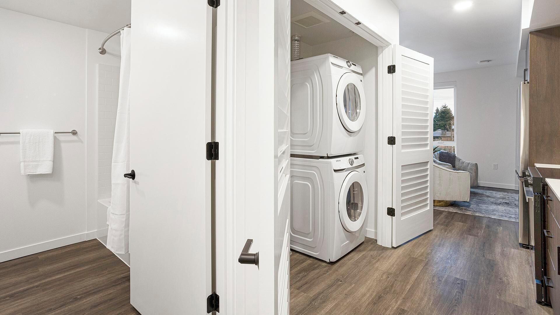 A modern laundry room with stacked washing machines, a wooden floor, and a view of the outdoors through an open door.