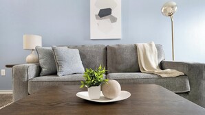 Una sala moderna con un sofá gris, cojines decorativos, una pequeña planta en maceta y una mesa de centro redonda de madera.