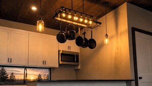 A modern kitchen with white cabinets, a stainless steel microwave, hanging pots, and two black stools. - Designer Lake Cabin- PickleBall, HotTub, Slide, EV Charging, Ice Baths, Biking (Dent)