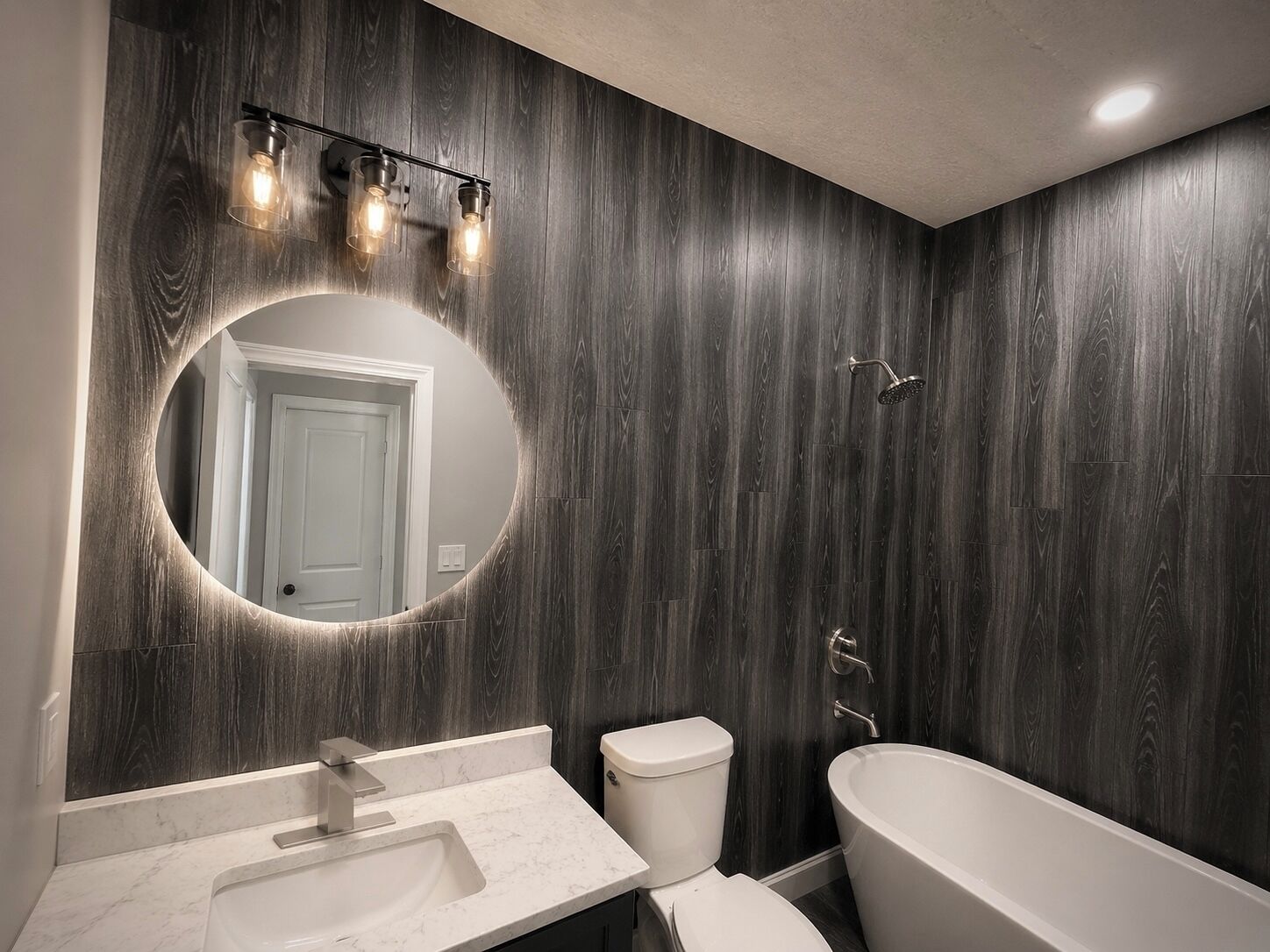 A modern bathroom with a dark wood panel wall, a round mirror, a white sink, a toilet, and a freestanding bathtub.