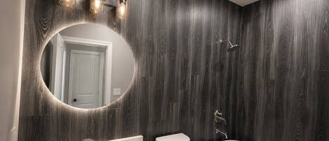 A modern bathroom with a dark wood panel wall, a round mirror, a white sink, a toilet, and a freestanding bathtub.