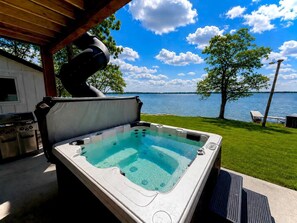 A hot tub with a view of a lake, a grill, and a covered patio area.