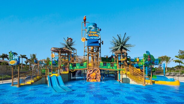 Un parc aquatique doté d’un grand toboggan, d’une tour à jeux d’eau et de palmiers.