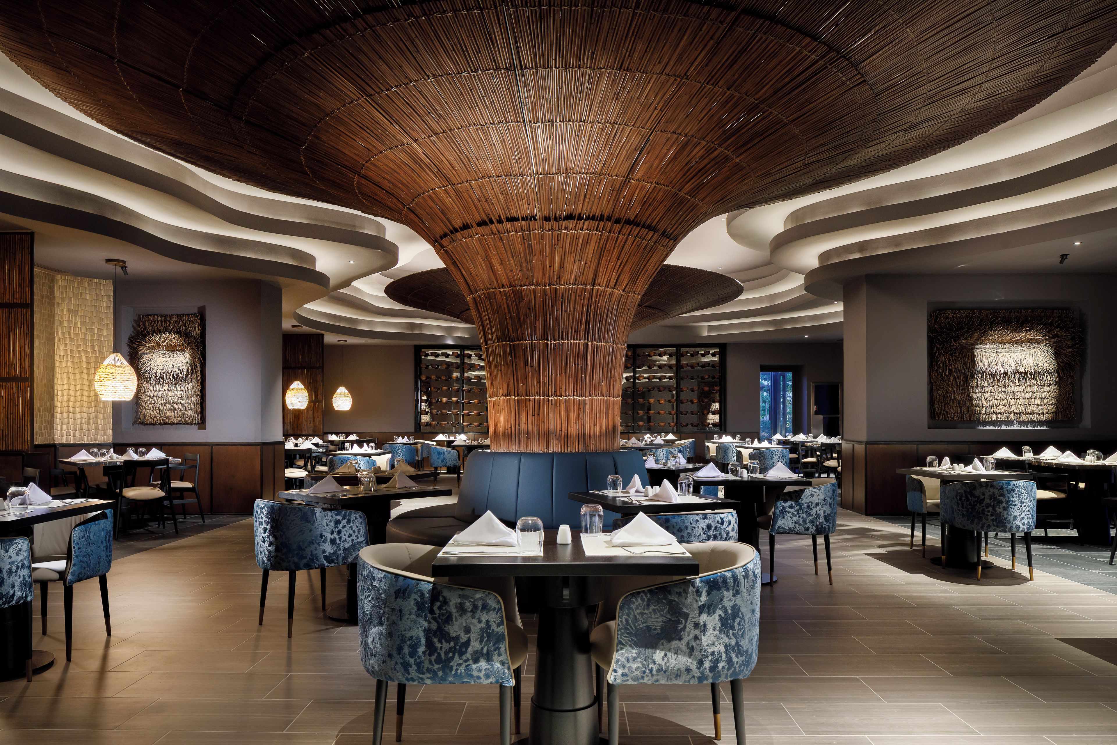 Un interior de restaurante con mesas redondas, sillas tapizadas en azul y un diseño único de techo de bambú.