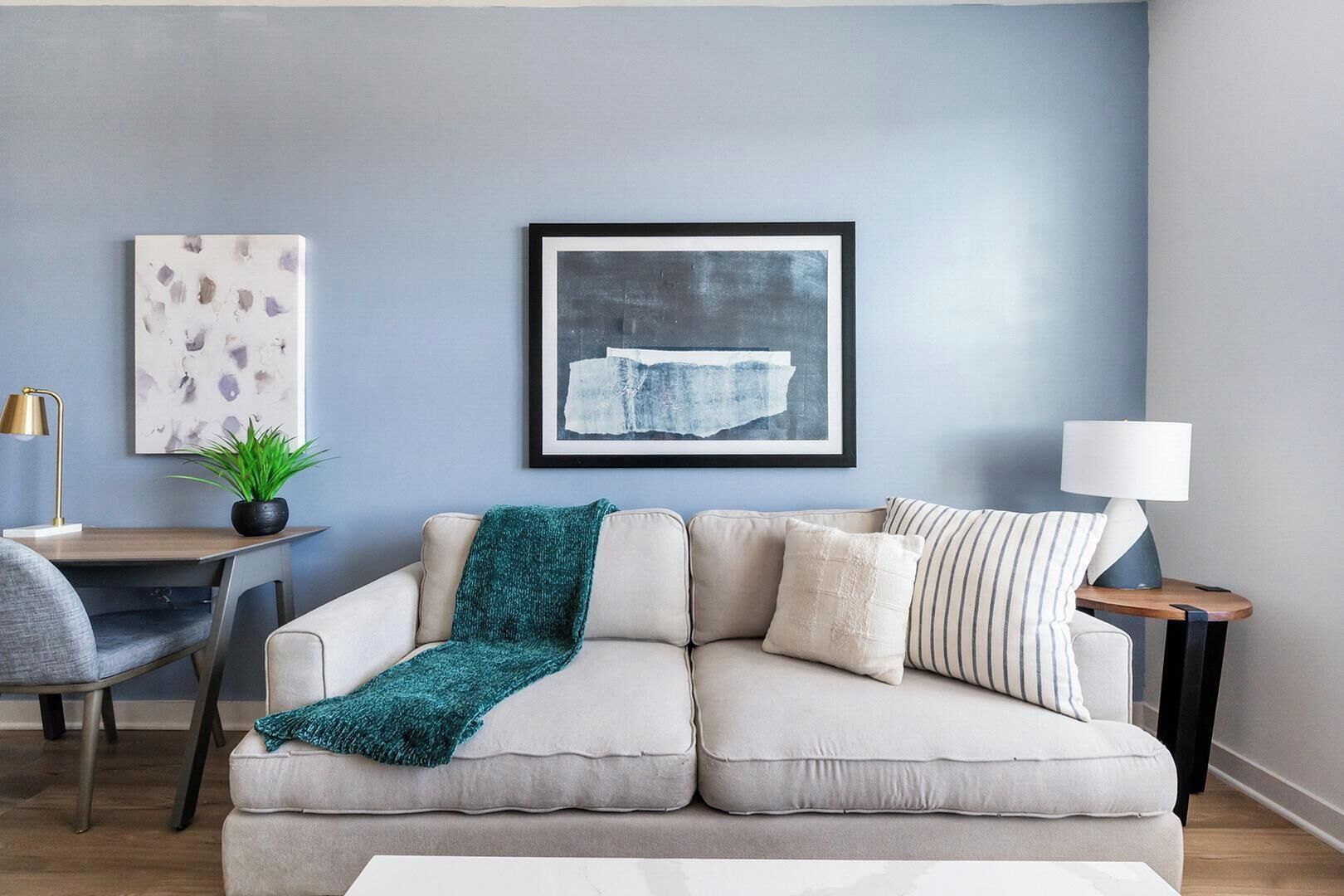 Ein Wohnzimmer mit einer Couch, einem Schreibtisch, einer Lampe und zwei gerahmten Kunstwerken an der Wand.