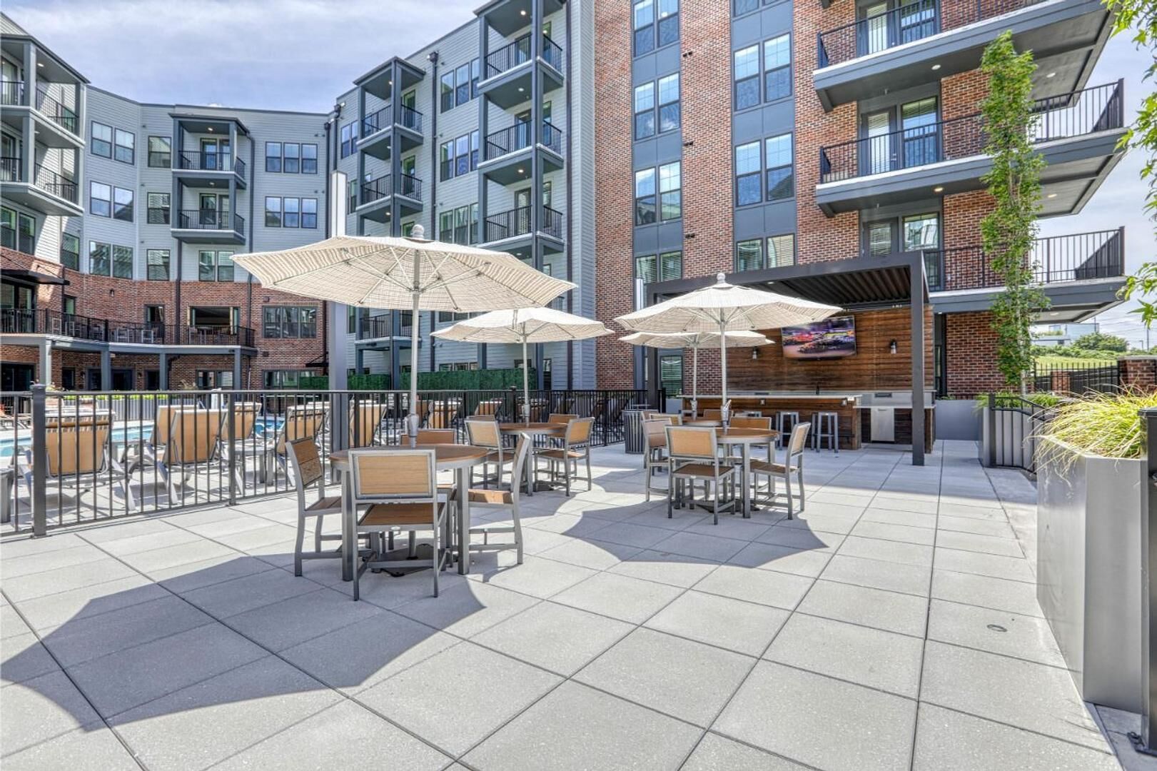An outdoor patio area with tables and chairs, umbrellas, and a pool in the background.