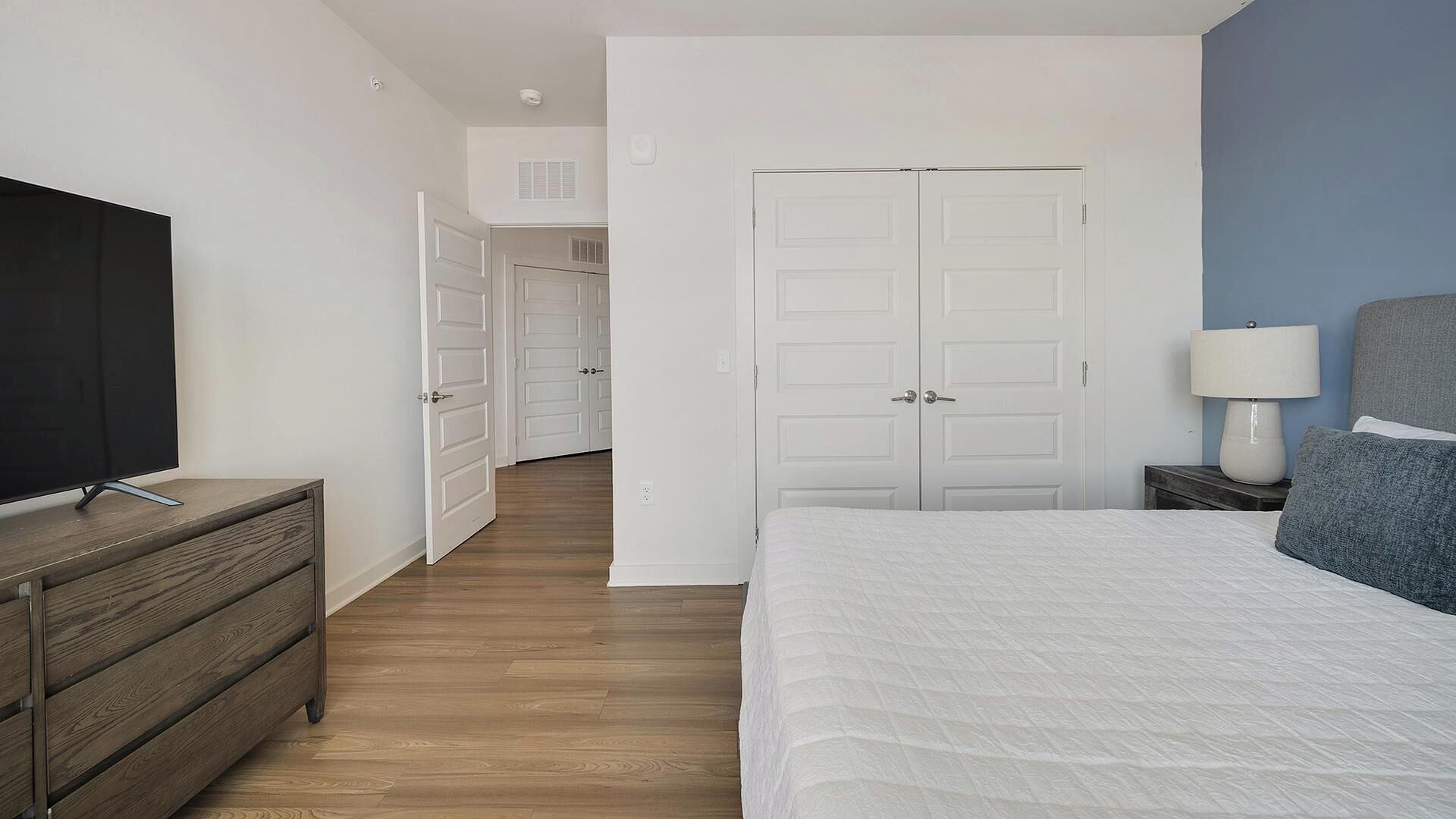 Een slaapkamer met een bed, een televisie op een dressoir, een nachtkastje met een lamp, en een deur die naar een andere kamer leidt.