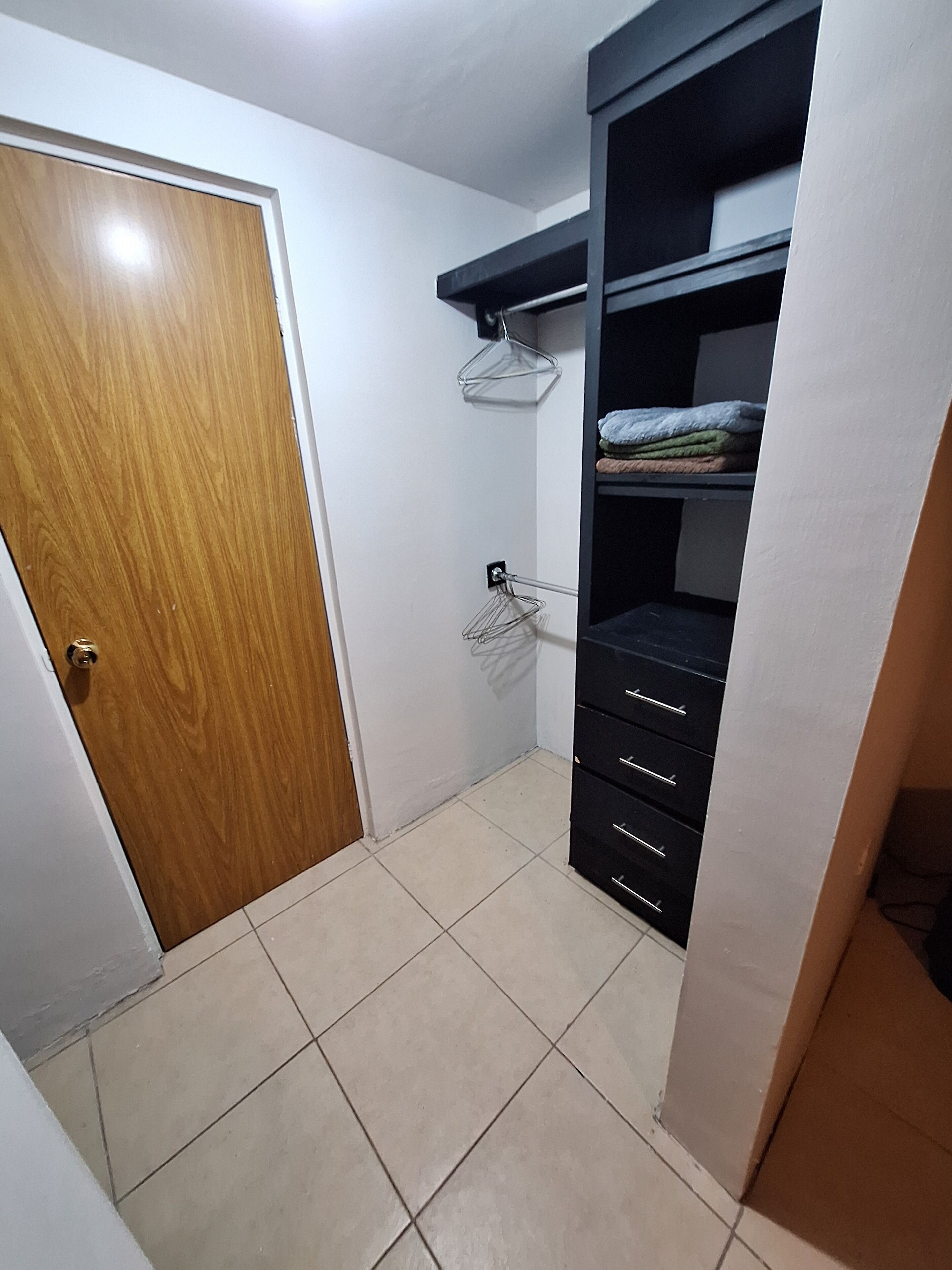 A room with a wooden door, tiled floor, a black wardrobe with drawers, and a metal hanger on the wall.
