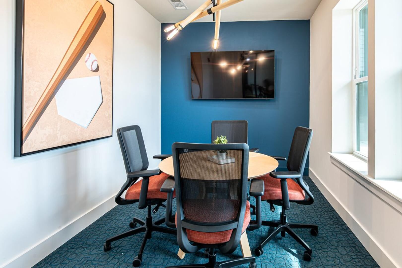 A conference room with a round table, four chairs, a large painting, and a TV on the wall.
