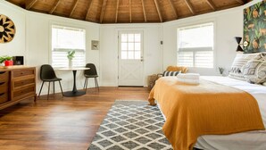 Ein Schlafzimmer mit Holzbalkendecke, einem Bett mit gelber Decke, einem Schreibtisch mit zwei Stühlen und einem Fenster mit Jalousien.