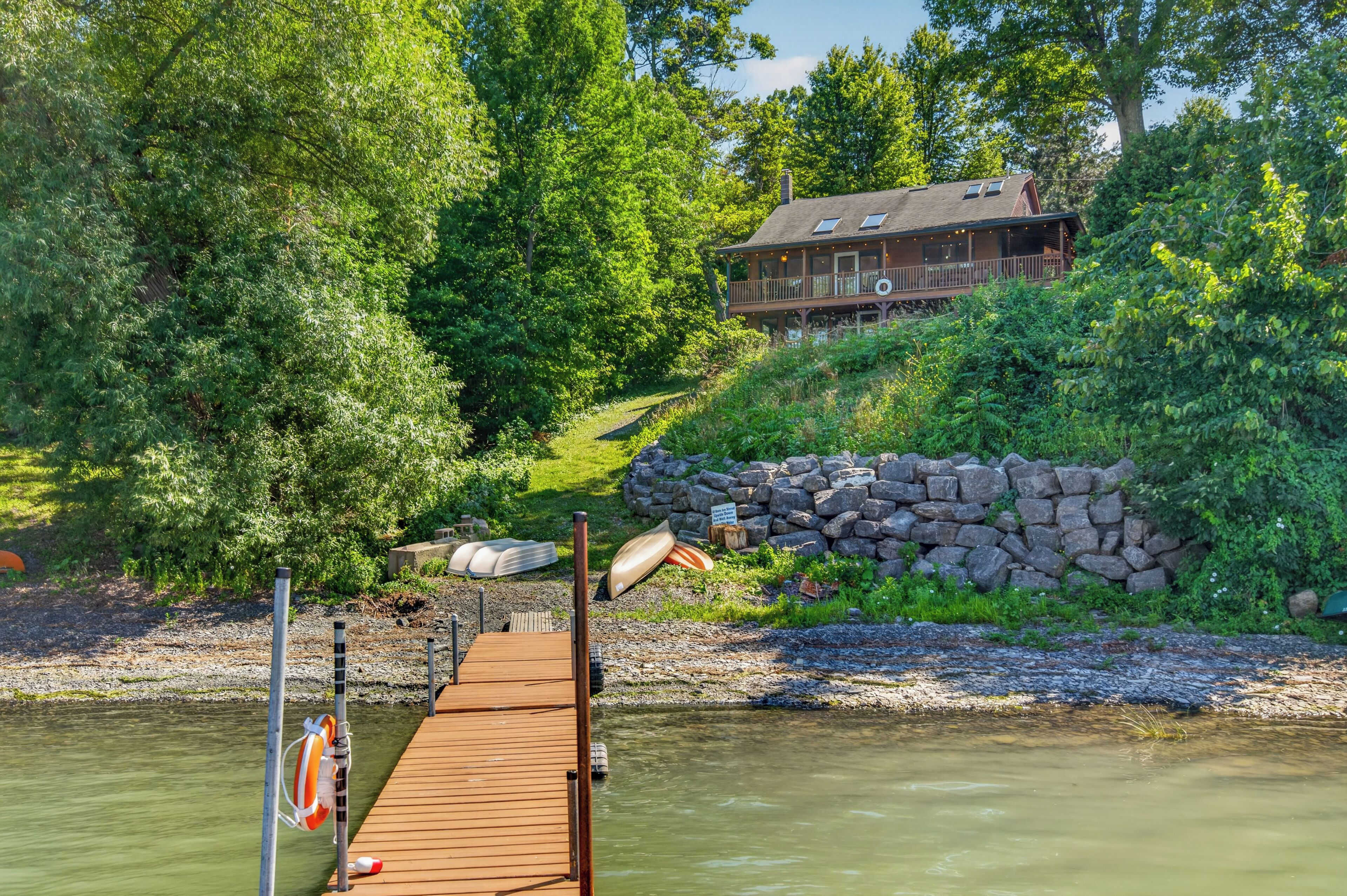 A wooden dock leading to a house surrounded by dense greenery and a stone wall.