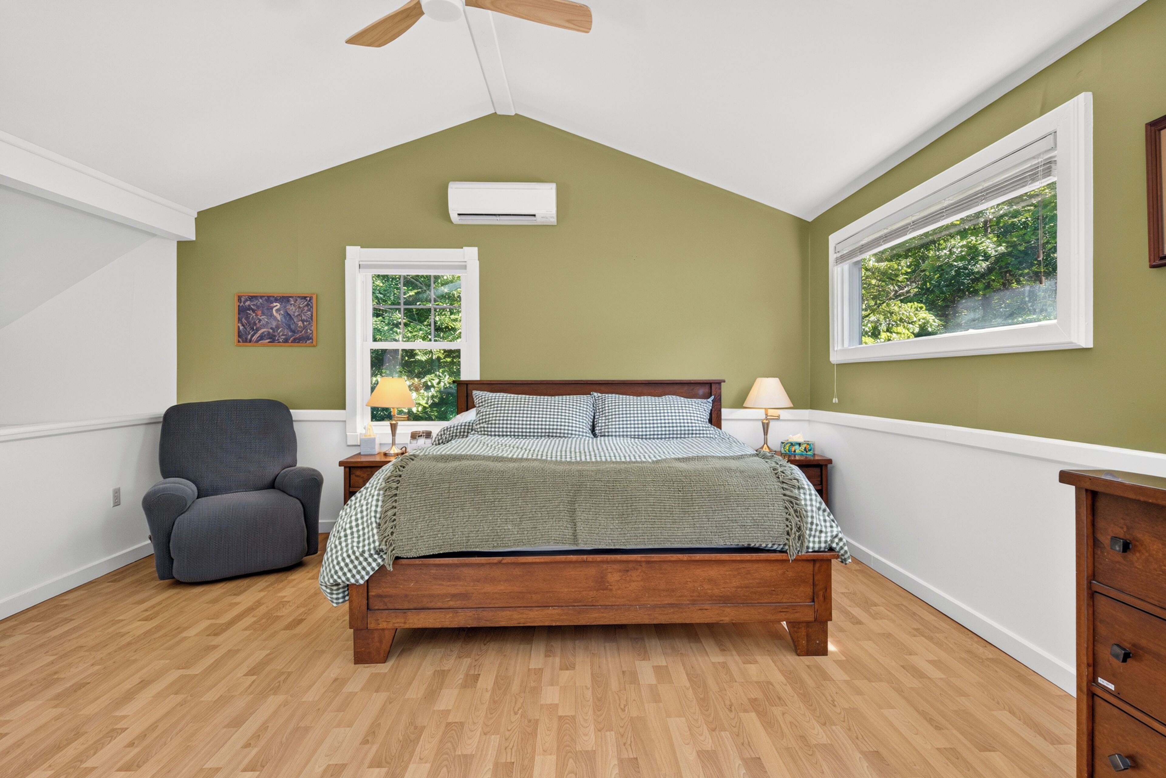Un dormitorio con una cama de madera, un sillón gris, una mesita de noche con una lámpara y un ventanal con vistas a la vegetación.