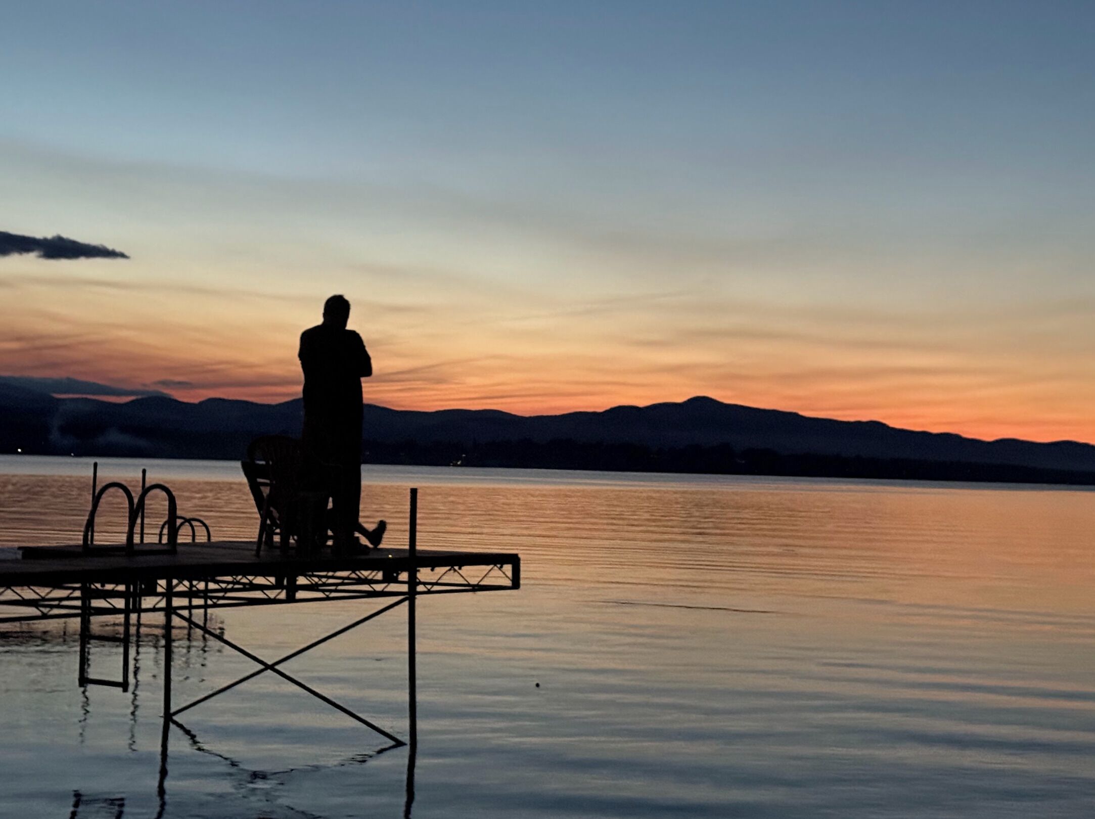 Una persona de pie en un muelle con una silla, contemplando un cuerpo de agua tranquilo y montañas distantes al atardecer.
