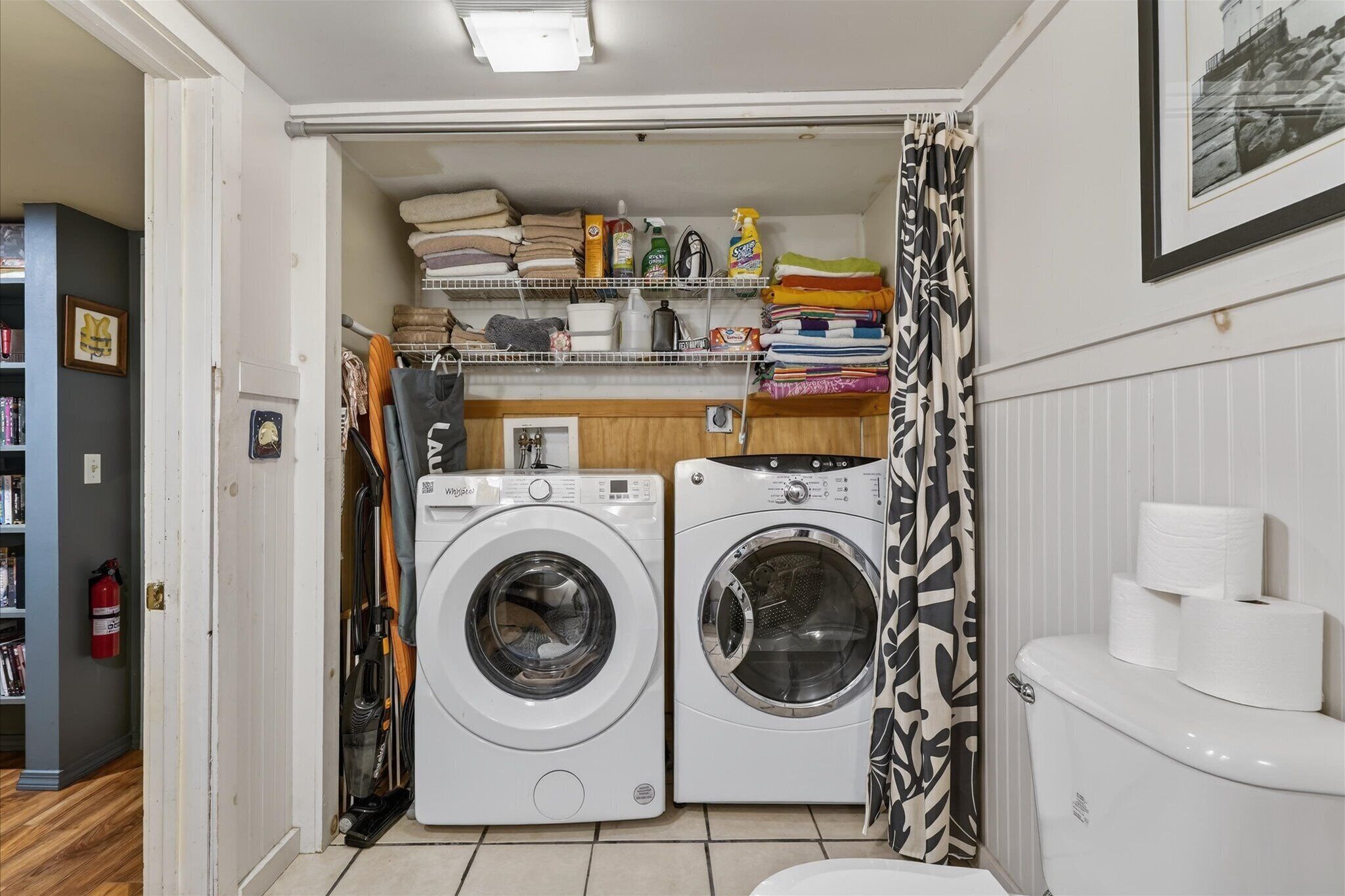 Un cuarto de lavandería con dos lavadoras, estantes con productos de limpieza y un inodoro con papel higiénico.