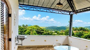 A balcony with a table and chairs, a hanging light fixture, and a view of a green landscape with hills. - Chácara 3 Poderes Piscina - 60 pessoas (Guararema)