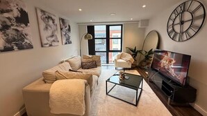 A modern living room with a sofa, coffee table, and TV. There are abstract paintings on the wall and a large clock. - Luxury 2-Bed Apartment | Gym | Jewellery Quarter (Birmingham)