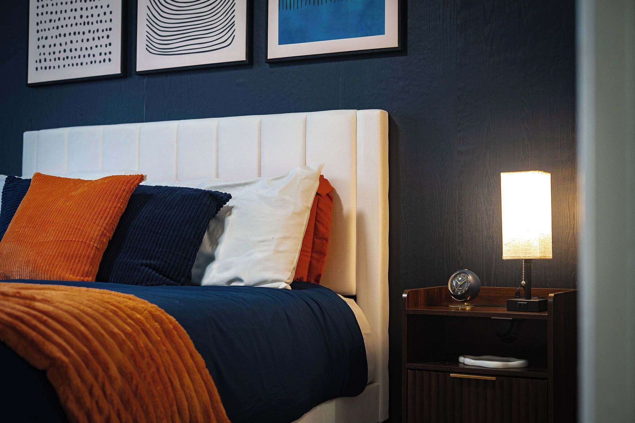 A neatly made bed with a white headboard, multiple pillows in various colors, a bedside table with a lamp, and a clock.