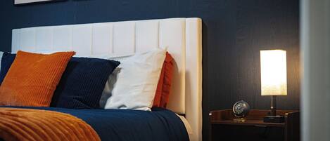 A neatly made bed with a white headboard, multiple pillows in various colors, a bedside table with a lamp, and a clock.