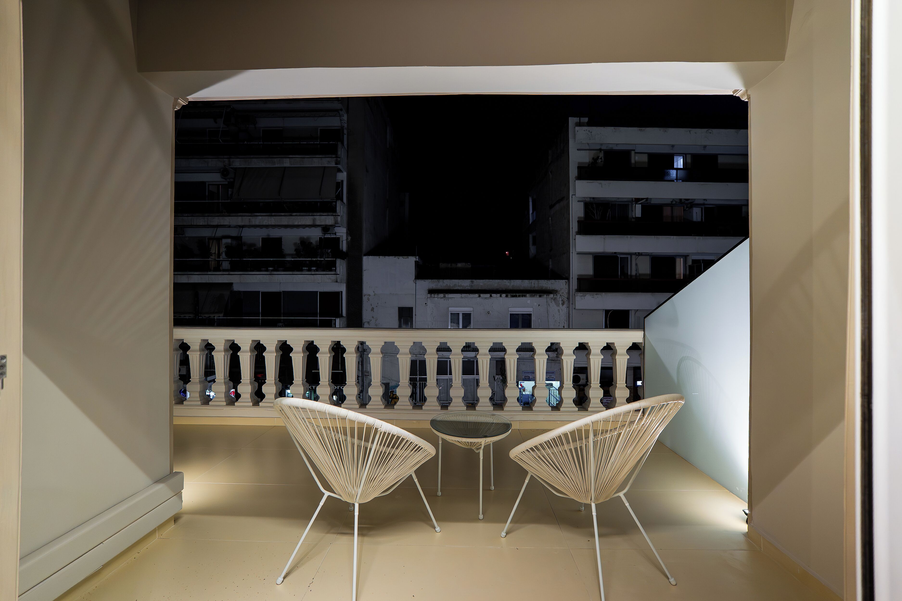 Uma varanda com duas cadeiras de arame brancas e uma pequena mesa, com vista para um horizonte urbano noturno com edifícios e uma escadaria.