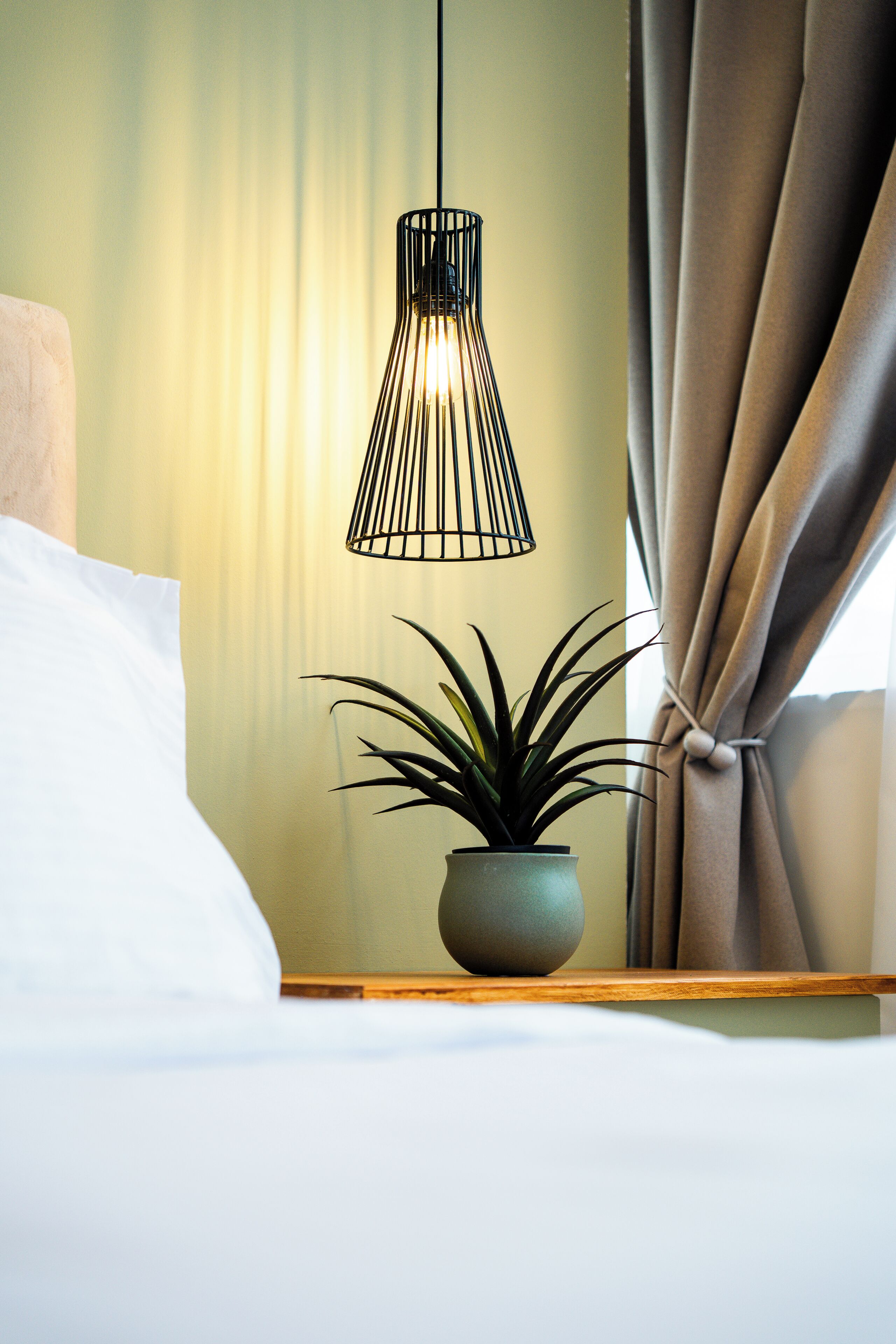 A neatly made bed with a white pillow, a potted plant on a wooden nightstand, and a hanging light fixture.