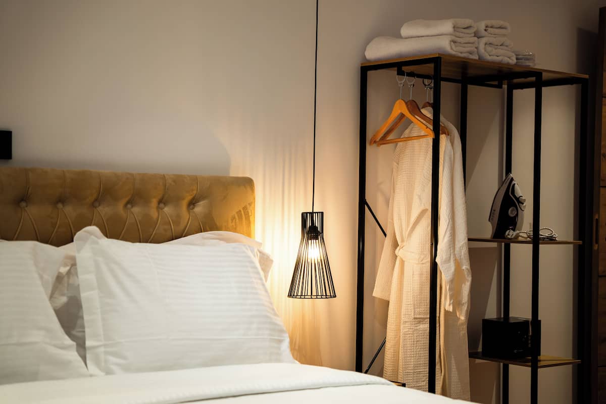 Cama bem arrumada com lençóis brancos, luminária pendente e prateleira de madeira com toalhas dobradas e roupão.