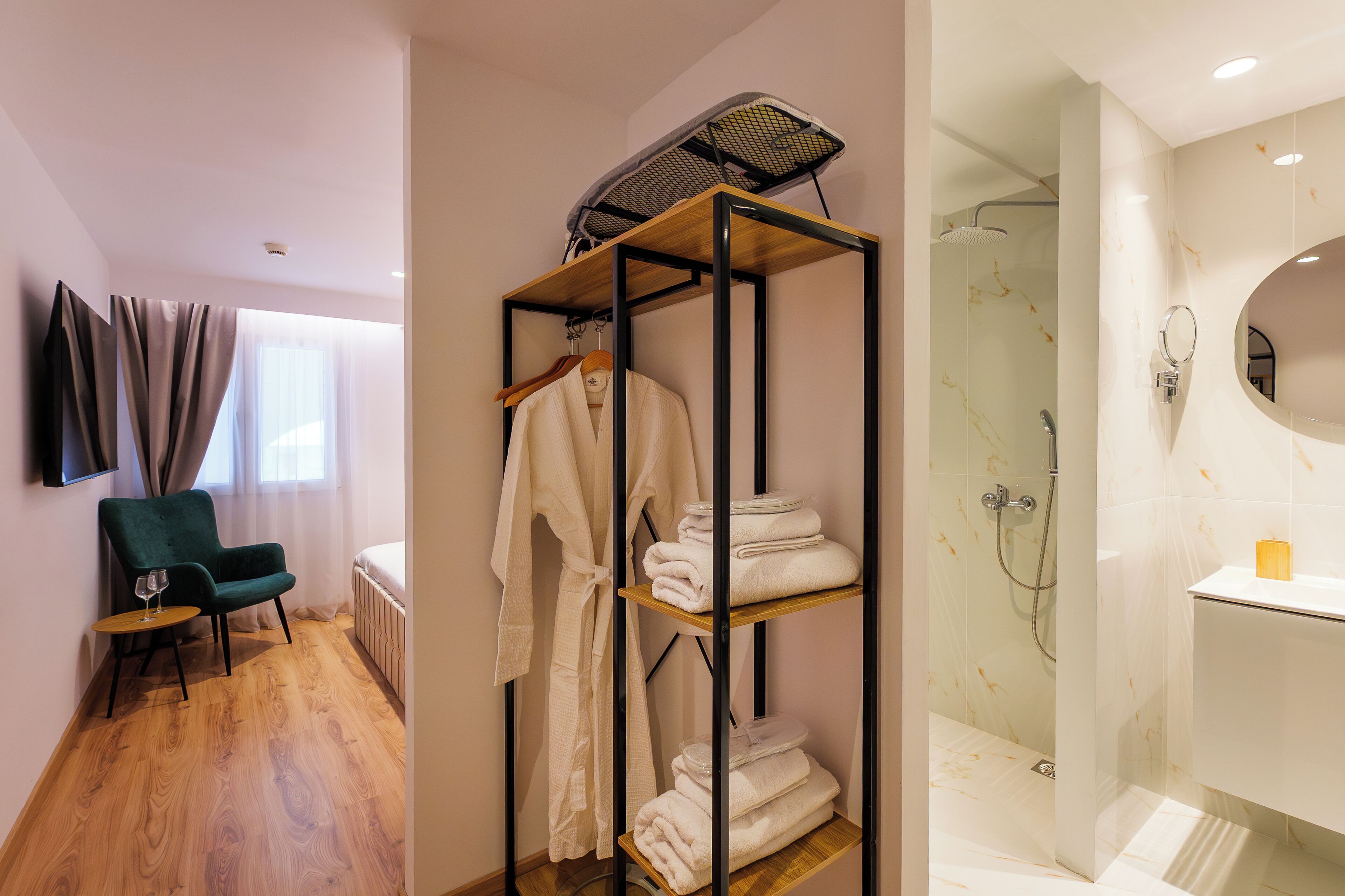 A hotel room with a wooden floor, a green chair, a shelf with folded towels, a robe, and a shower area with a glass door.
