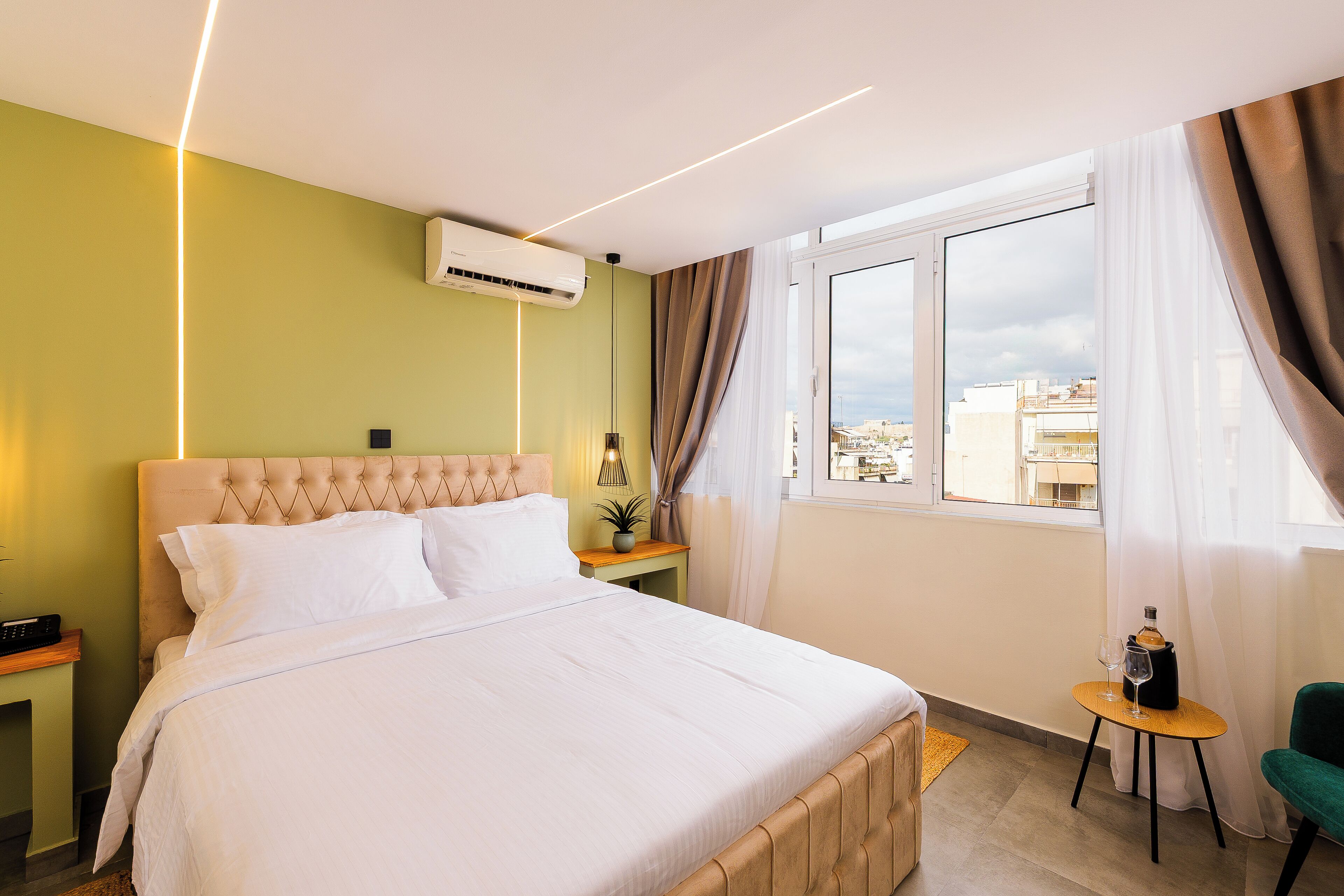A hotel room with a bed, a small table with a bottle and glass, a chair, a nightstand, and a window with curtains.