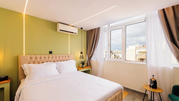 Habitación de hotel con cama, una mesita pequeña con una botella y un vaso, una silla, una mesita de noche y un ventanal con cortinas.
