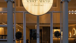The entrance of Aether Athens Urban Hotel with a prominent illuminated sign, classical columns, and a modern interior visible through the glass doors.